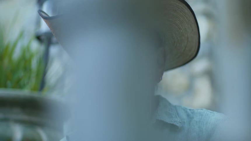 A closeup shot of a mans face wearing a straw hat shows him looking directly forward while his head is partially framed by vertical streaks of outoffocus light and a green plant in the background