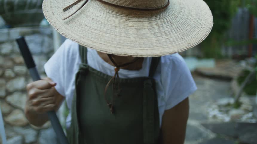 A woman wearing a widebrimmed straw hat and green overalls over a white shirt stands outdoors holding the black handle of a garden tool and looking down as she works