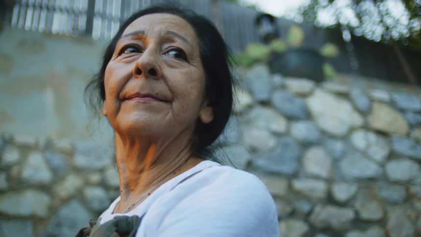 An elderly woman with dark hair looks off to the side outdoors by a stone wall appearing calm