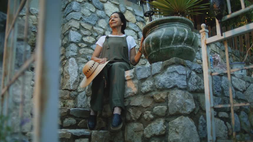 Woman sits on a stone staircase holding a hat and admiring the garden
