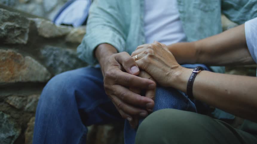 Two people sit sidebyside with one persons hand resting on the other persons hand placed upon the mans knees