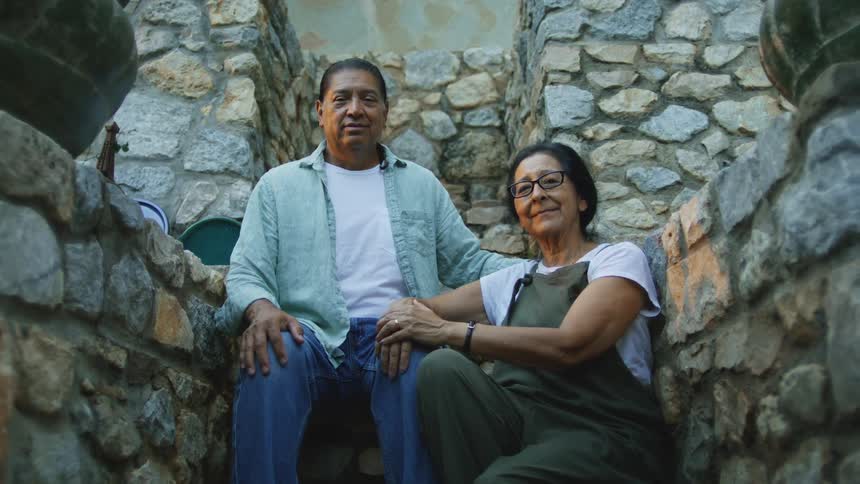 A man and a woman sit closely on a stone staircase framed by rough stone walls with the mans arm around the womans shoulders and the couple looking forward smiling