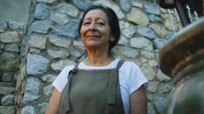 An elderly woman in an apron smiles warmly while standing in front of a stone wall
