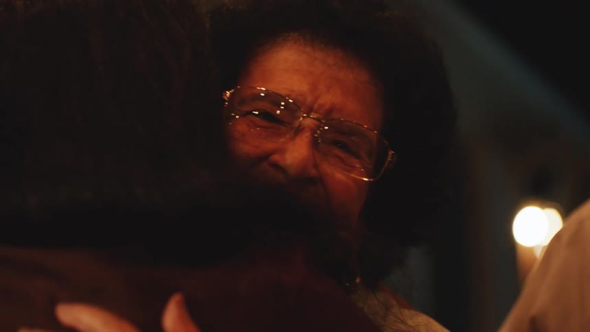 The video is a warm closeup shot of an elderly woman with an afro and goldrimmed glasses appearing to cry with emotion while embracing another person her expression conveys deep feeling in a dimly lit interior setting