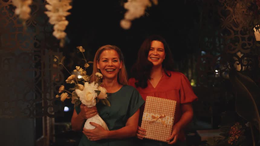 Two smiling women walk through an archway with flowers and a gift at night