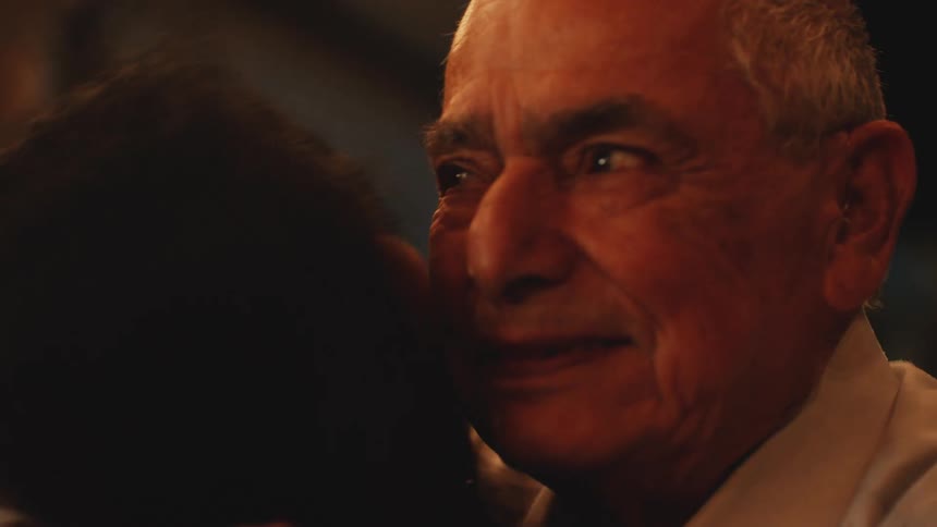 An elderly man with a heartfelt expression hugs and is kissed on the cheek by a woman whose head is mostly off screen