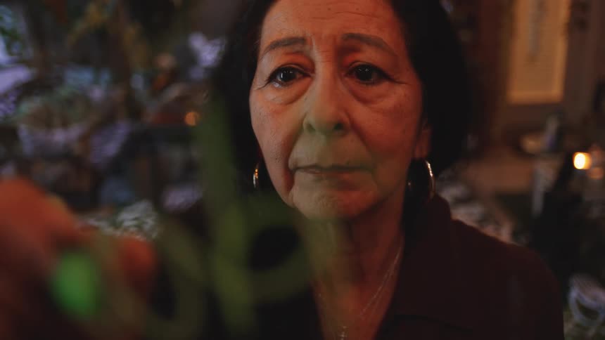 An elderly woman adorned with makeup and earrings writes on a board with a pen in a welllit room