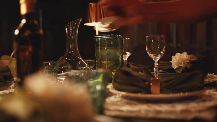 A person reaches across an elegant dinner table to set up wine glasses under warm ambient light