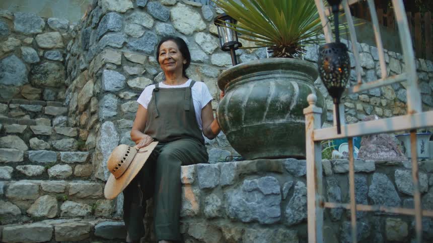 An older woman sits on a stone wall next to a large potted plant and a metal archway holding a straw hat in her lap and smiling directly at the camera