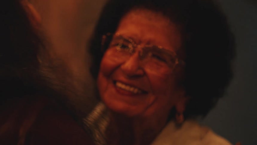 Elderly woman wearing glasses smiles emotionally while hugging someone