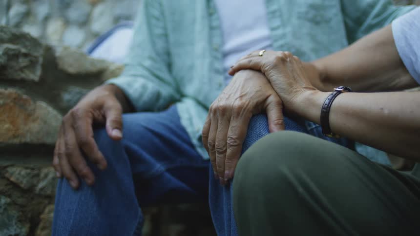 An elderly man sits outdoors at midday gently supported by another person whose handadorned with a ring and a watch
