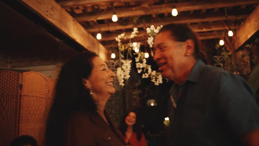 Older man and woman kiss on the porch at night while their friends and family watch and smile