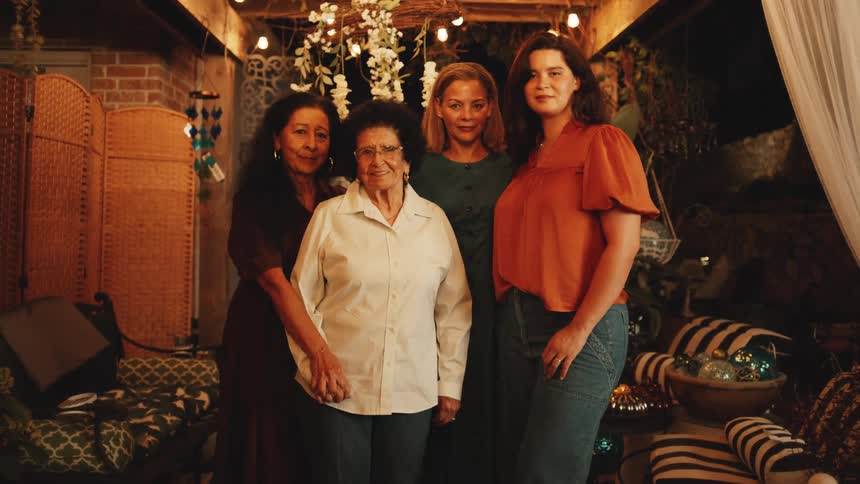 Four women of different ages stand together and smile at the camera in a warmly lit bohemian inspired patio decorated with white string lights and potted plants at night