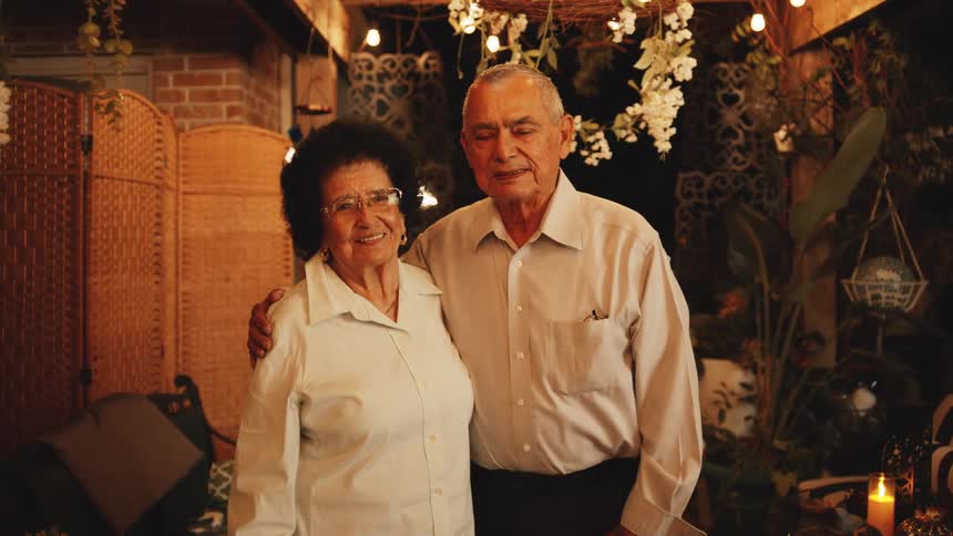 Elderly couple smiles side by side in a cozy setting decorated with hanging lights and flowers