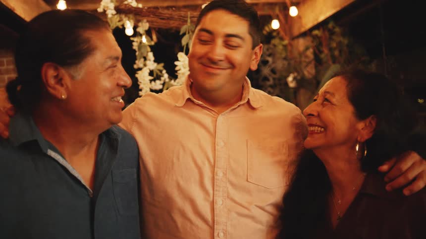 A man stands between an older man and an older woman with their arms around his shoulders and all three look at each other and smile warmly under overhead string lights