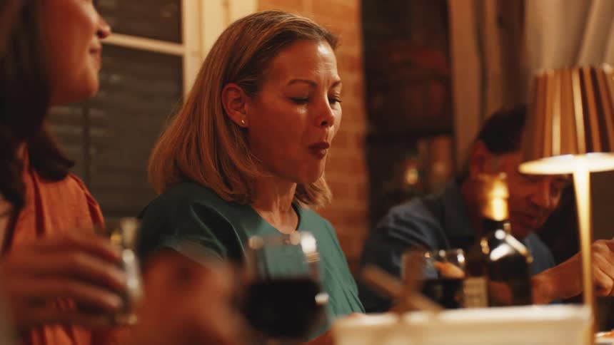 A woman with blond hair wearing a green shirt sits at a table and eats food from a fork then speaks to another person while a man and a lamp are visible in the background