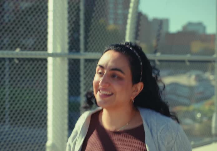 A woman with dark hair stands on a metalfenced pedestrian bridge in a city smiling broadly and reaching up to push her hair back from her face