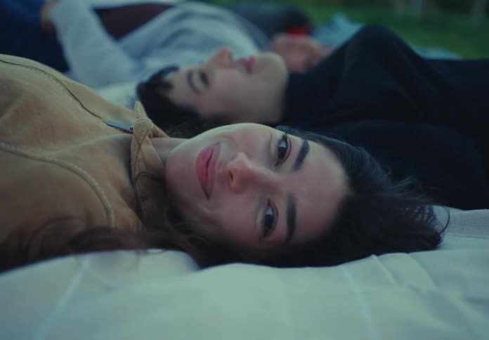 Three young female friends lie side by side on a blanket spread across a grassy lawn outdoors smiling warmly as they enjoy each others company
