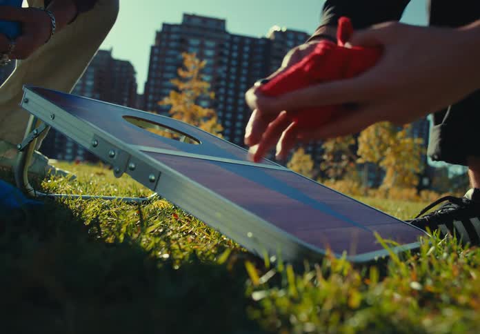 Hands quickly grab and remove two red bean bags resting on a board pitched on a grassy lawn with large apartment buildings and autumn trees visible in the sunlit urban park background