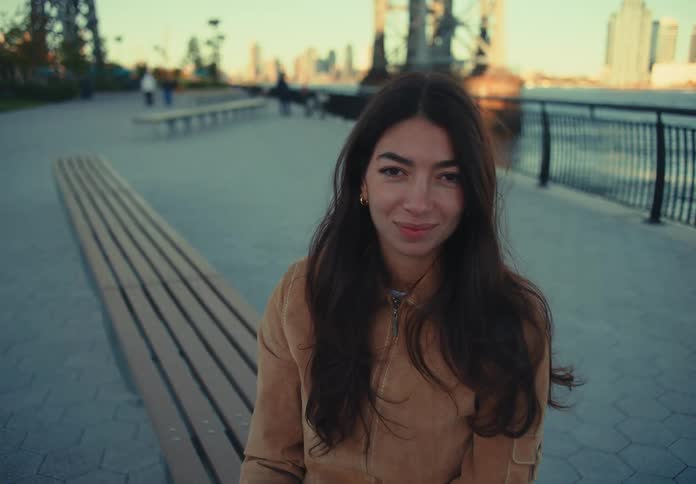 A woman sits on a park bench smiling with the city skyline behind
