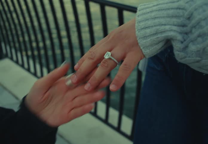 One persons hand reaches out and places a large diamond ring onto the finger of another persons hand which is resting near a metal railing overlooking water