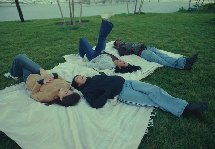 Four people lie down on their backs on a white blanket spread over a grassy lawn looking up and talking with one person briefly lifting a leg into the air before resting it again