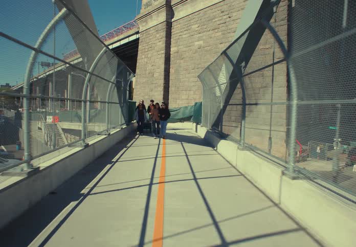 A dolly in shot shows a group of casually dressed young people walking and chatting on a sunny bridge walkway enclosed by a chain link fence overlooking a highway and urban buildings