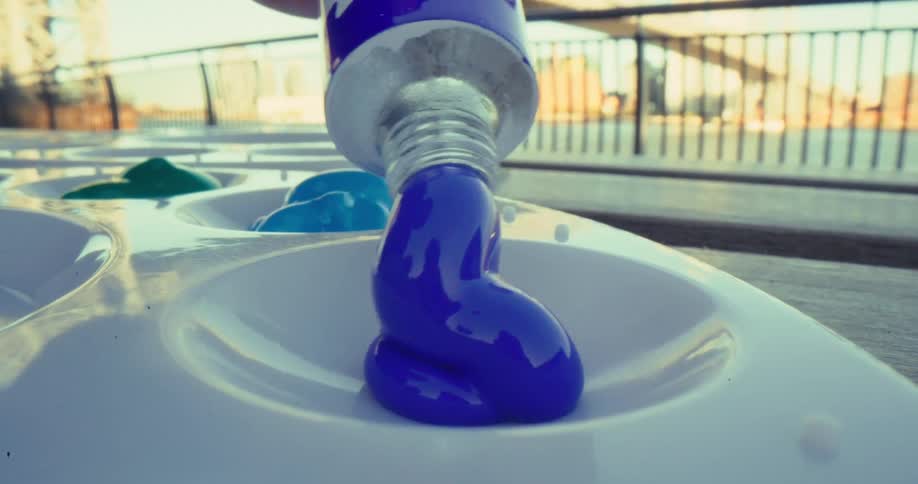 A close view shows thick blue paint being squeezed from a tube onto a white plastic palette with green and light blue paint already present set against a sunny outdoor background with an urban railing