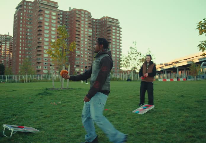 A couple of friends have a picnic in a public park at midday as a young man stands up from the ground and walks to another young man the two greeting each other with a handshake