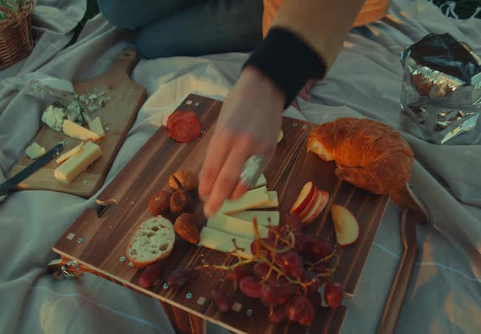 A person reaches for red grapes on a small wooden picnic table set with cheese bread and a croissant on a blanket