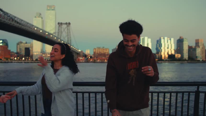 A couple of college friends stand beside a city river at midday laughing and enjoying themselves as they shake hands and congratulate a nearby couple with cheerful smiles