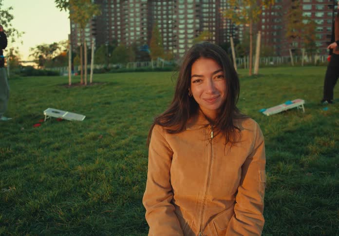 A woman sitting on a blanket on a grassy lawn looks over her shoulder then turns to face the camera and smiles with apartment buildings and two men playing a beanbag toss game visible in the park background