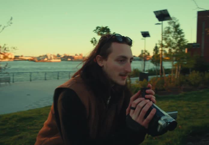 A long dolly shot of a young man with long dark hair smiling and filming with a vintage video camera in a grassy park with a large suspension bridge and a city skyline visible behind him at sunset
