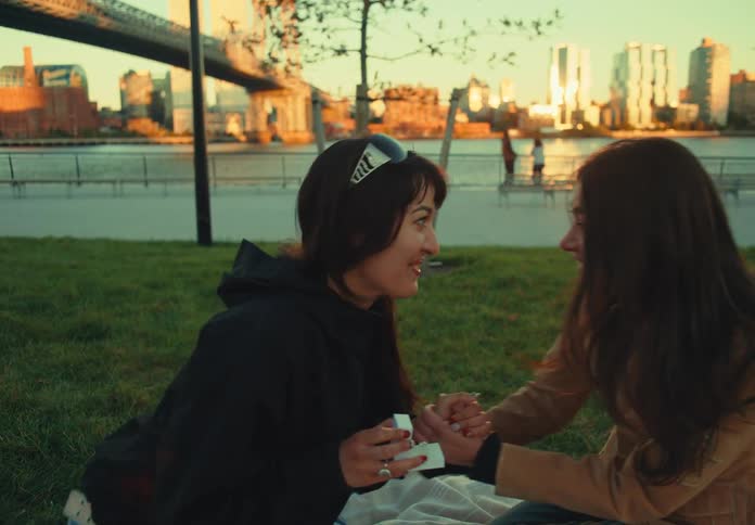 Two woman sit facing each other on a blanket on a grassy lawn in an urban park where one of them opens a small box causing the other person to raise a hand to her mouth in surprise before the first person stands up and walks away