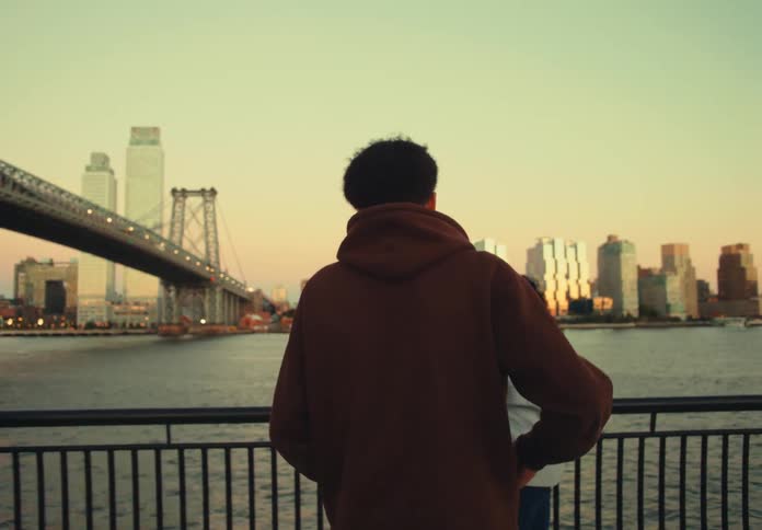 Two friends stand by a railing fence beside a river chatting casually a young man approaches one of the young women then kneels to propose sparking shock and excitement from her behind them the city skyline and a bridge stretch across the background