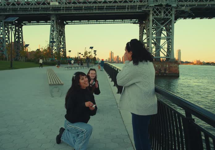 A woman walks along a paved waterfront promenade with a city skyline in the background kneels down to open a box and propose to another woman who reacts with surprise and joy before the two women embrace and hug affectionately