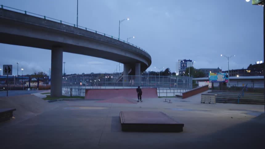 A person in dark clothing rides a skateboard across a paved skatepark containing ramps and platforms passing under a large concrete overpass structure in an urban area at twilight