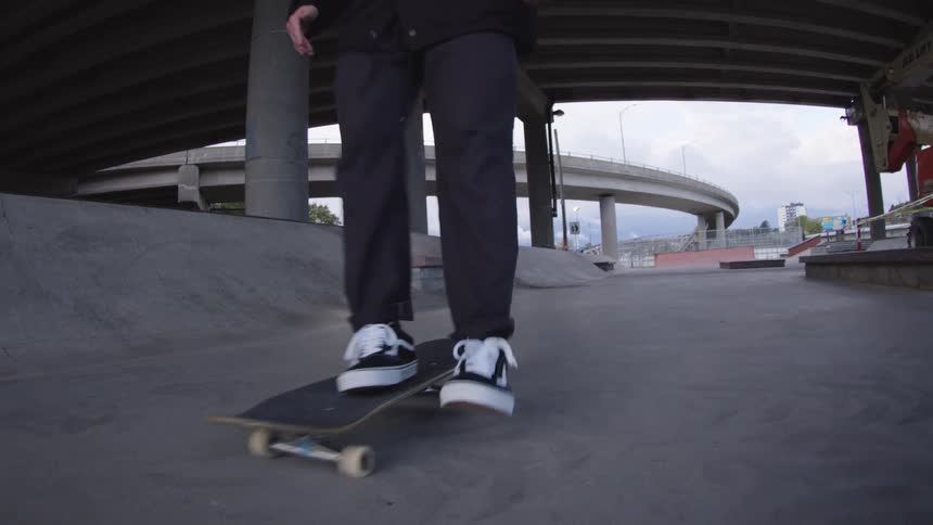 The video is a lowangle shot of a person skateboarding on flat ground under a bridge or overpass at an urban skate park the skater performs a small trick while being filmed closely from the front with the concrete pillars and overpass structu