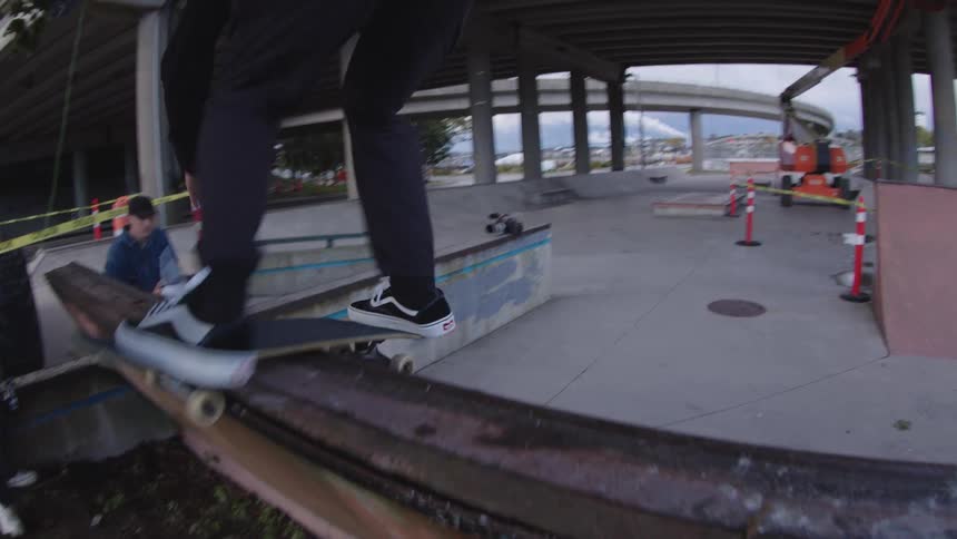 A fisheye view shows a man wearing a black suit and hat grinding a rusty metal rail on a skateboard in an urban skate park beneath a concrete bridge overpass
