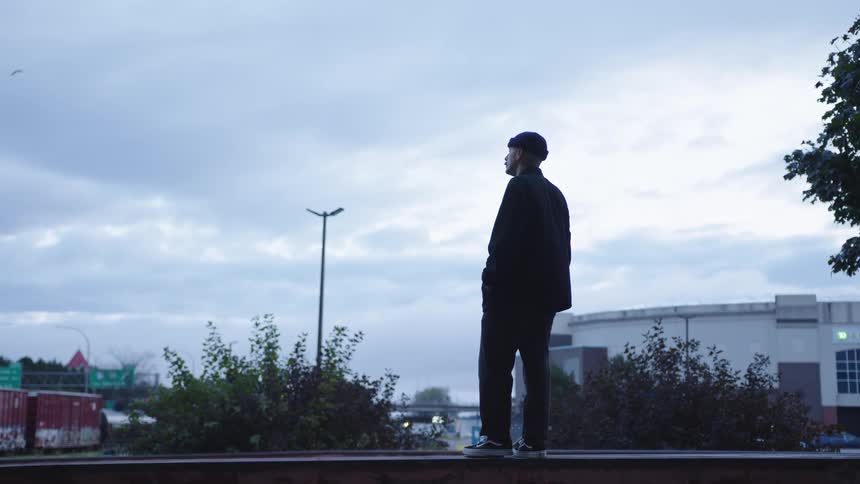 The video shows a man standing on a ledge overlooking a mostly cloudy suburban industrial area with a road and a large building in the background he is silhouetted against the pale sky hands in his pockets taking in the dreary view