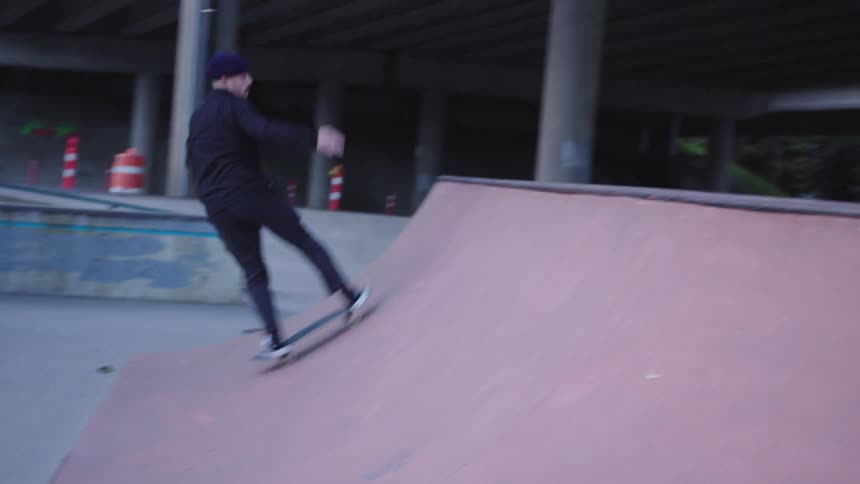 The video shows a skateboarder dressed in all black riding a halfpipe at an outdoor skate park beneath a bridge or overpass the person skates smoothly up and down the ramp successfully performing a turn near the top edge of the structure