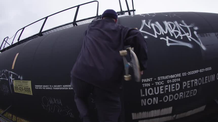 Male skateboarder climbs a train tanker car while holding a skateboard