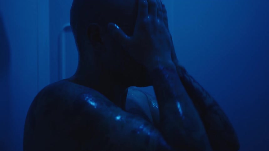 A close moody view shows a bald man with tattooed arms standing in a dark shower running his hands down his wet face and shoulders under intense blue lighting