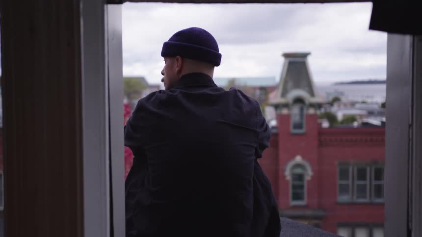 A person wearing a dark beanie and dark jacket sits with their back to the camera looking out a window opening at a city composed of red brick buildings under a gray sky