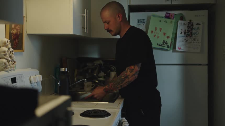 A bald man with a mustache and full arm tattoos wearing a black shirt washes dishes in a kitchen sink next to a stove and refrigerator in a dimly lit apartment