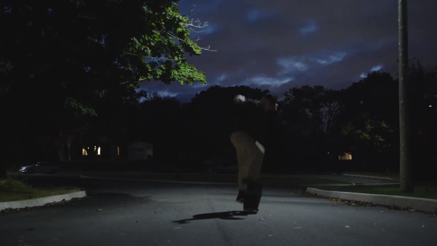 A man wearing dark clothing and khaki pants is seen from a low angle skateboarding down a dark empty neighborhood street at night under a cloudy dark sky with streetlights glowing faintly in the distance