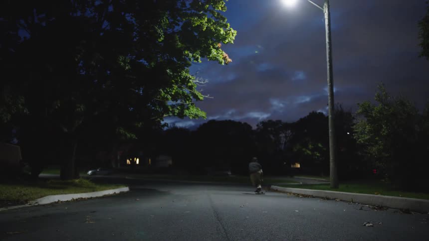 A person pushes a skateboard down a curved paved street in a residential area moving away from a bright streetlight on the right side of the road under a dark cloudy sky
