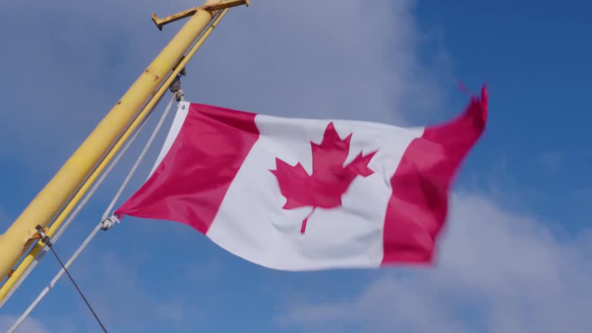 A low angle close view shows the canadian flag flying proudly from a yellow mast against a bright blue sky with scattered white clouds