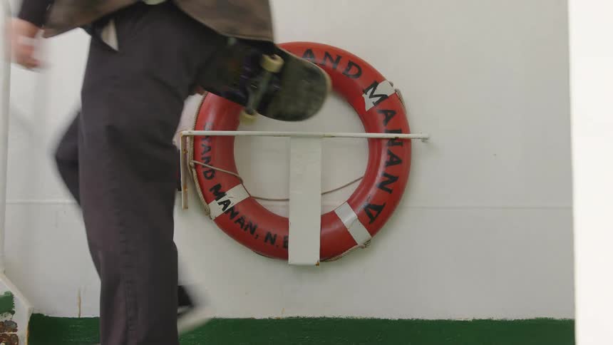A person walks quickly from right to left across the frame a round orange life preserver is mounted on a white wall a green stripe runs along the bottom of the wall and he is carrying a skateboard