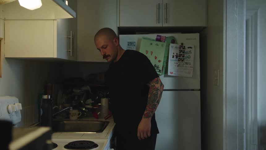 Man with shaved head washes pan in compact kitchen with white cabinets and warm lighting